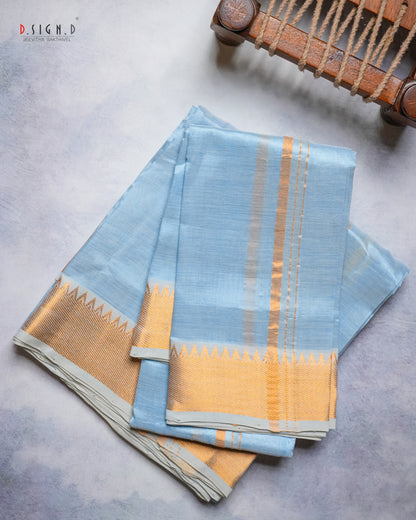 A folded light blue silk dhoti & vastram with a wide gold zari border and vertical gold stripes, displayed on a textured gray surface with a wooden loom visible in the top right corner.