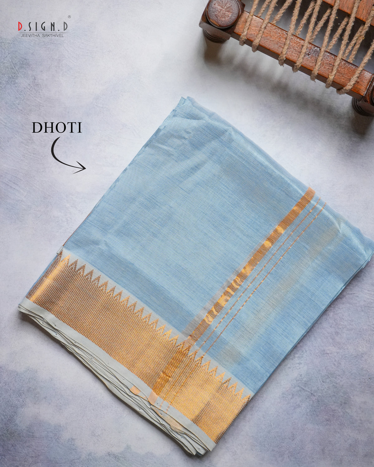 A folded light blue pure silk dhoti with a wide gold zari border and decorative gold motifs, displayed on a textured gray surface with a wooden loom prop in the corner. 