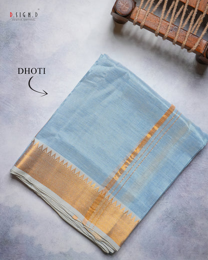 A folded light blue pure silk dhoti with a wide gold zari border and decorative gold motifs, displayed on a textured gray surface with a wooden loom prop in the corner. 