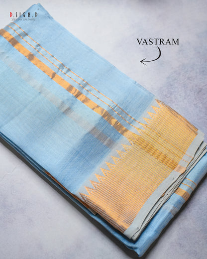 A folded light blue silk vastram with a wide gold and silver zari border, displayed on a textured gray surface. 
