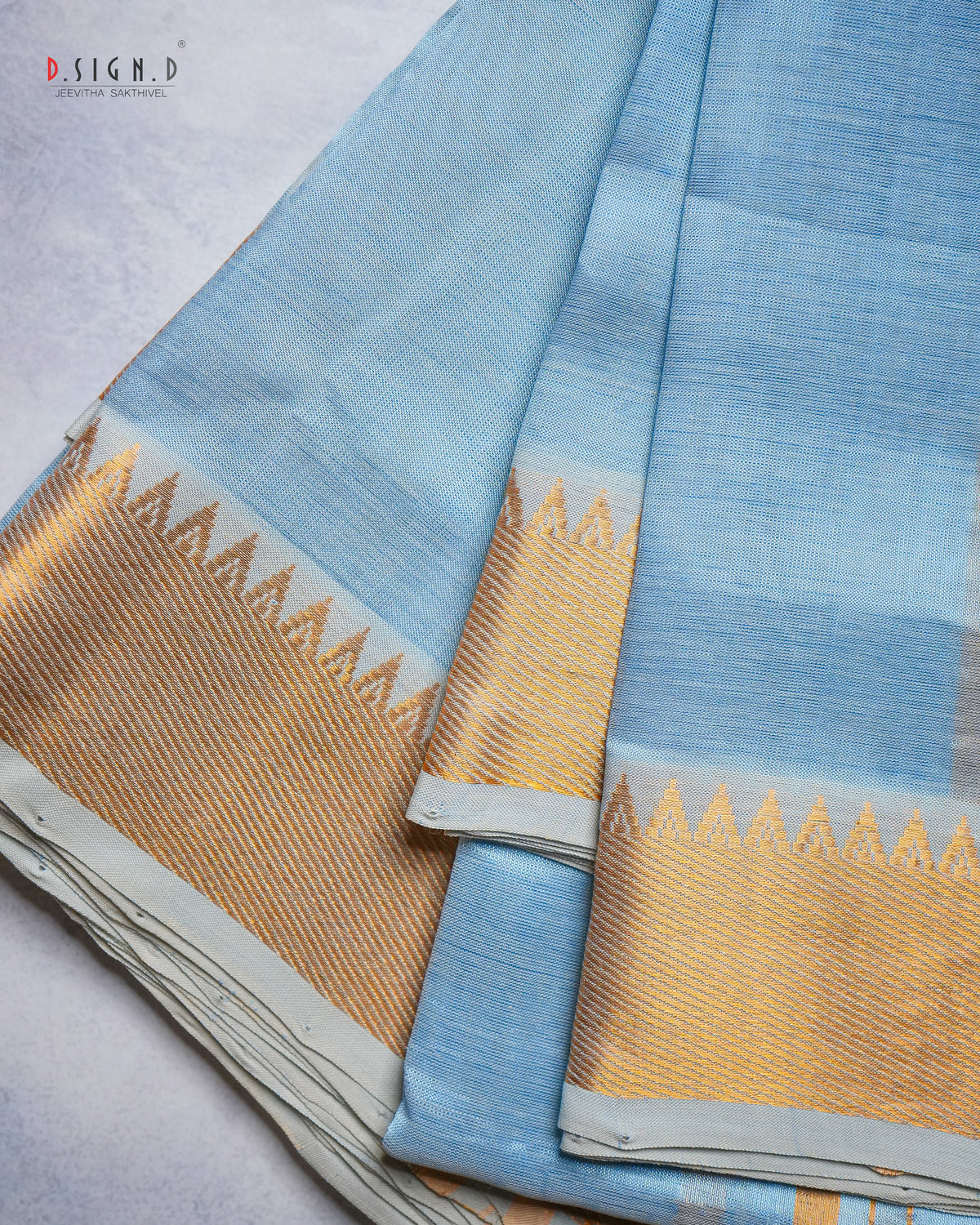 A folded light blue dhoti & vastram with a wide gold zari brocade border, displayed on a textured light gray surface. 