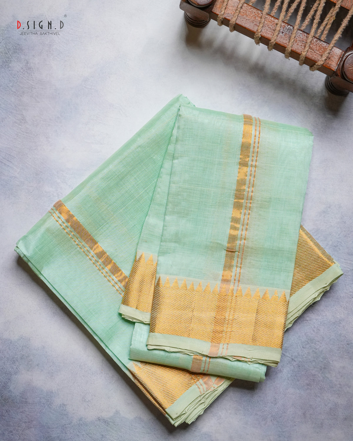 A folded mint green dhoti & vastram with a wide gold zari border and a small gold triangle motif, displayed on a textured gray surface with a wooden object partially visible in the top right corner.