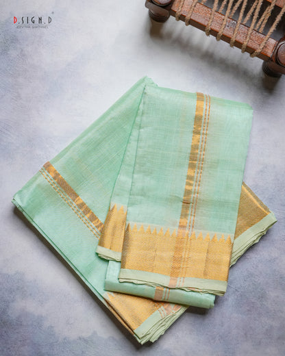 A folded mint green dhoti & vastram with a wide gold zari border and a small gold triangle motif, displayed on a textured gray surface with a wooden object partially visible in the top right corner.