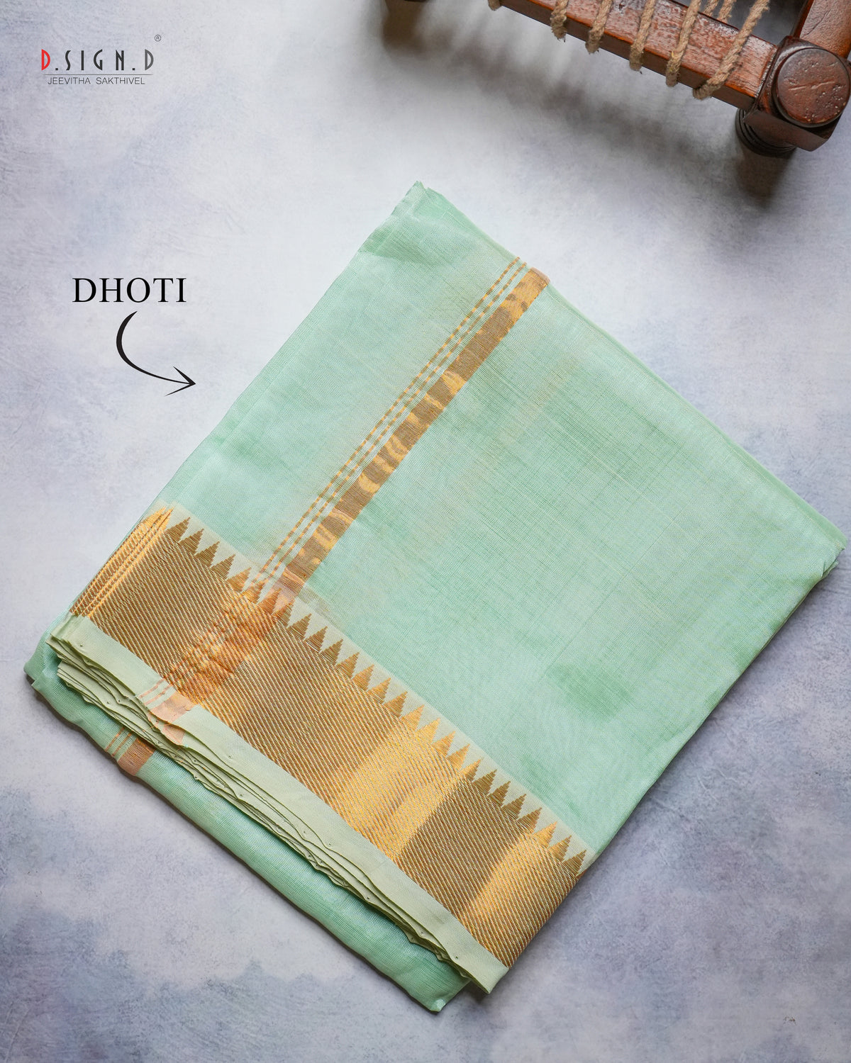 A folded mint green cotton dhoti with a gold zari border, displayed on a textured gray surface with a wooden object in the top right corner.