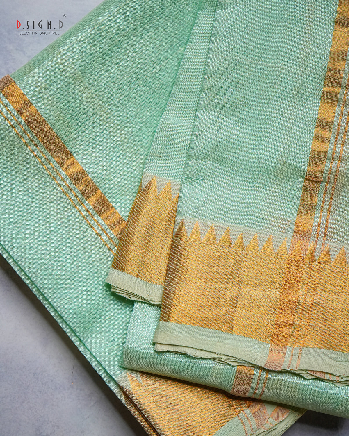 A folded mint green dhoti with a wide gold zari border and a decorative gold triangular patterned edge, displayed on a neutral gray surface.