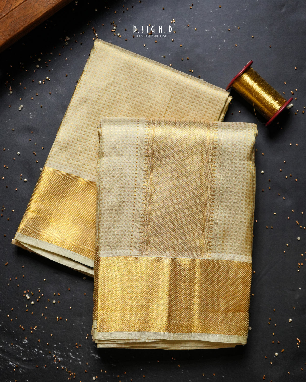 Two folded gold-colored silk dhoti or fabric pieces with a woven metallic border, arranged on a dark textured surface with a spool of gold thread nearby.