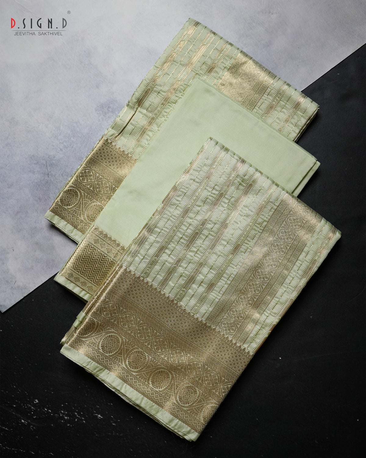 A folded light green and gold brocade fabric, likely a Dhoti or textile, displayed on a split gray and black background. The fabric features intricate metallic gold zari patterns, a detailed border, and a solid light green inner layer.