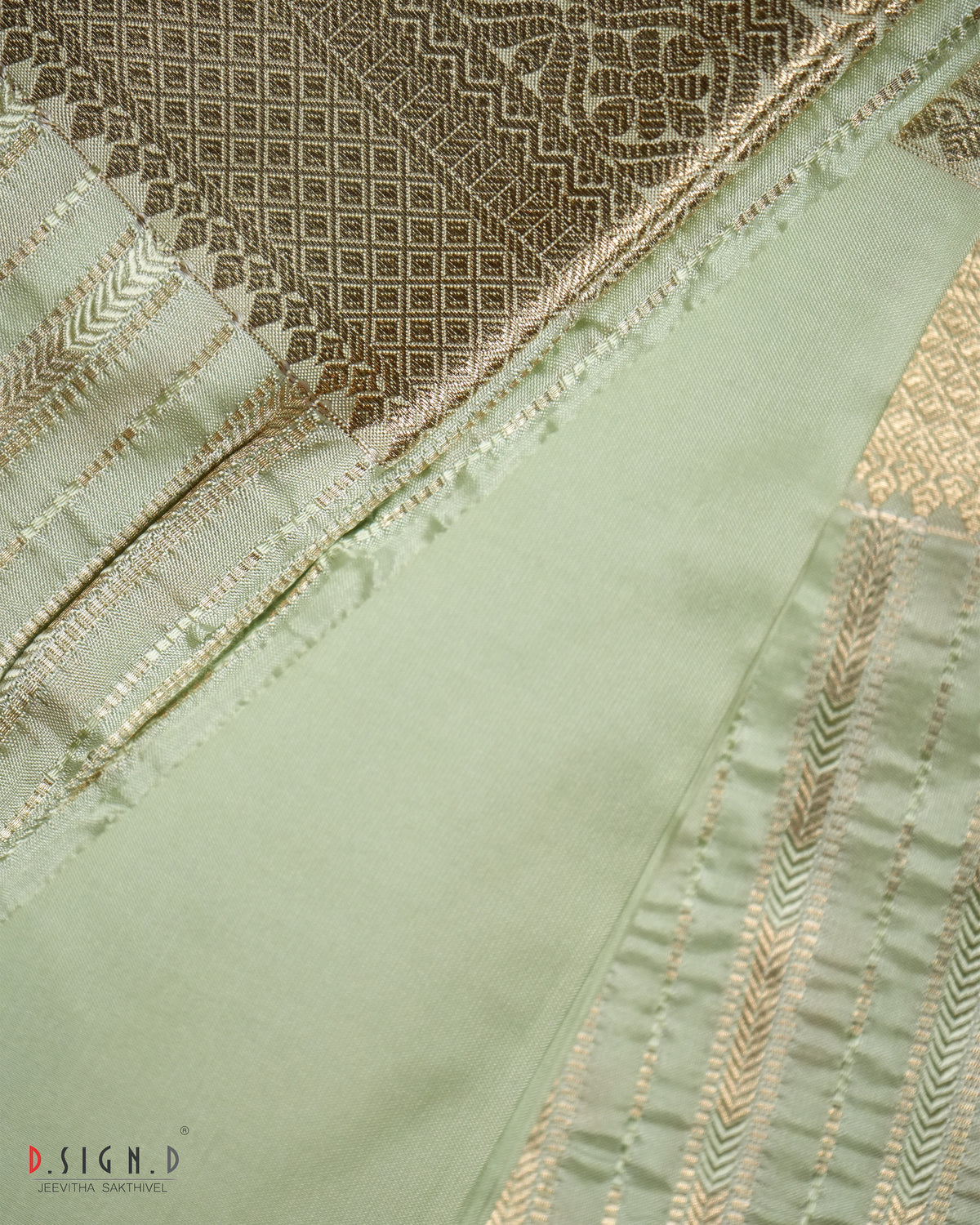 A close-up of a light sage green fabric with gold zari embroidery and a contrasting brown patterned brocade section, showing detailed stitching and texture.