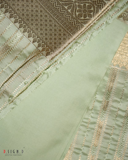 A close-up of a light sage green fabric with gold zari embroidery and a contrasting brown patterned brocade section, showing detailed stitching and texture.