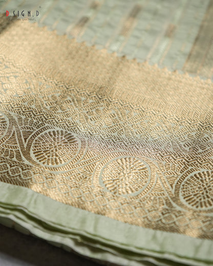 A close-up of a light sage green silk dhoti or fabric with a wide gold border featuring intricate geometric and circular woven patterns.
