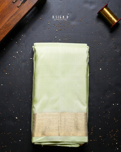 A folded light green silk dhoti set with a gold zari border, displayed on a dark textured background with decorative props.