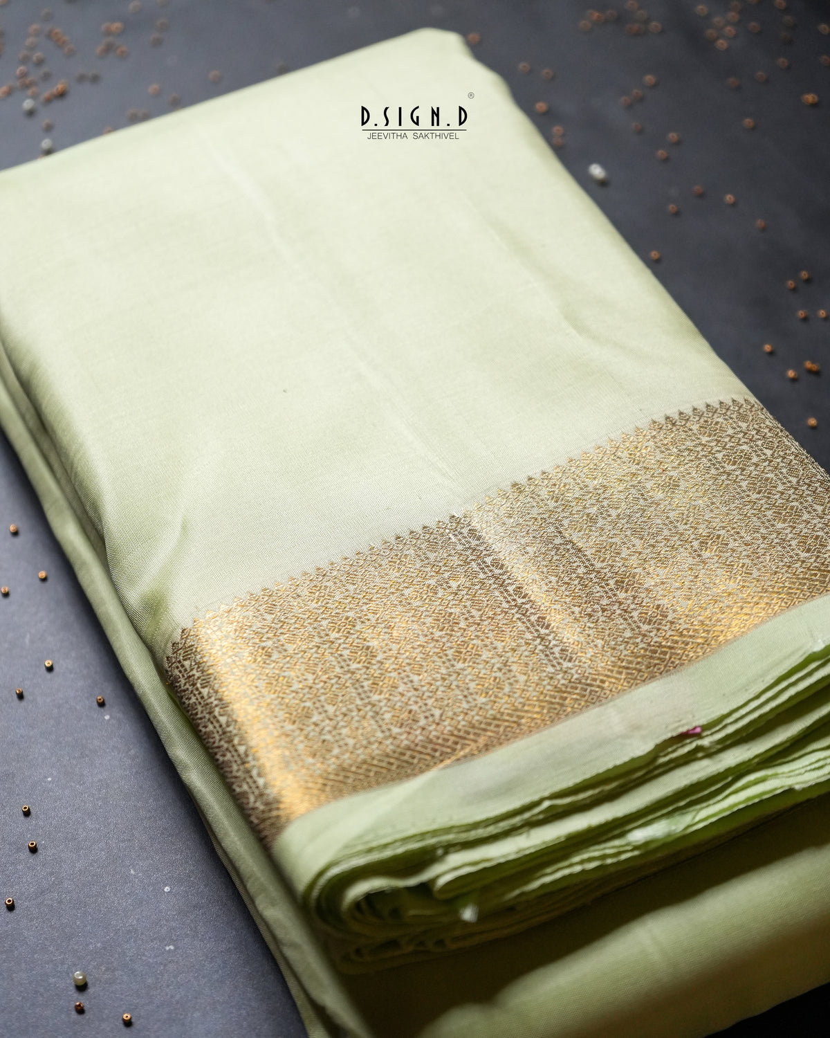 A folded light mint green dhoti set with a wide metallic gold zari border, displayed on a dark textured surface with scattered decorative seeds. 