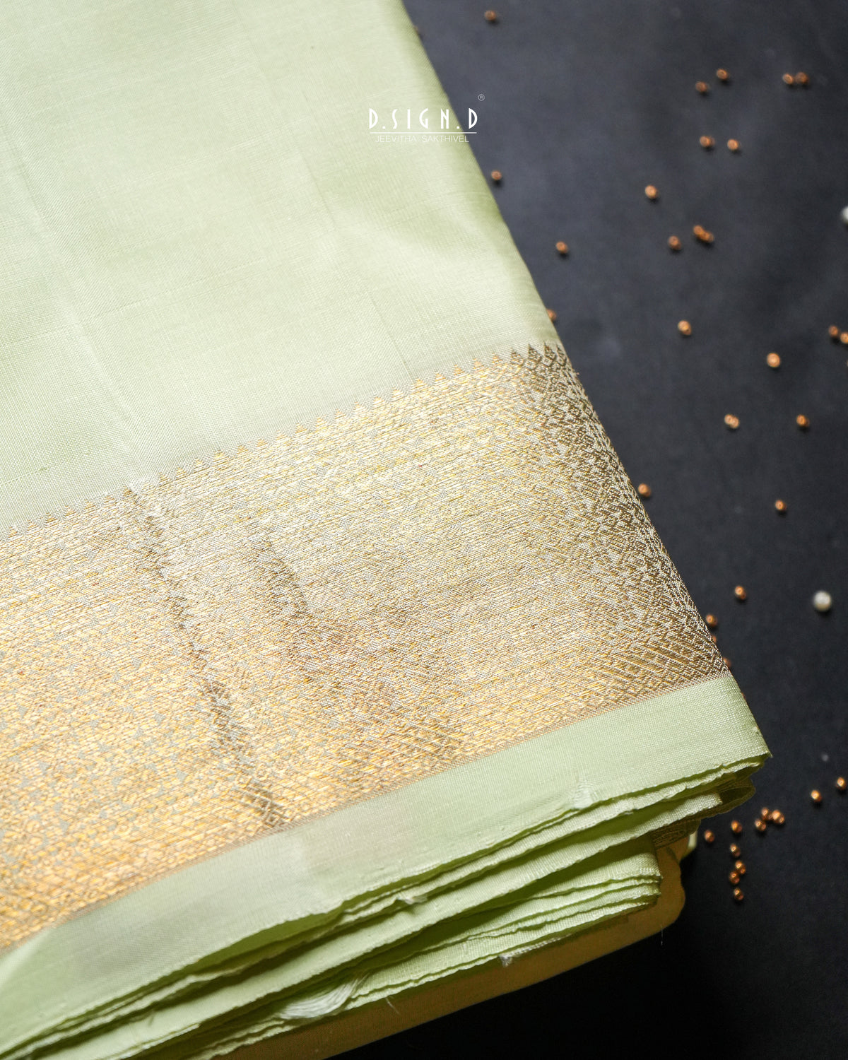 A folded light green dhoti or fabric piece with a wide metallic gold border, photographed on a dark background with scattered small seeds.