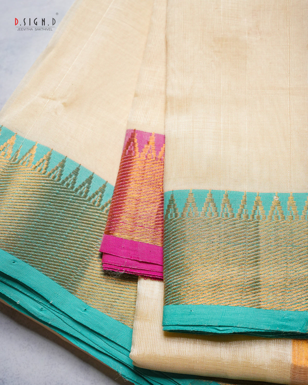 A folded cream-colored silk dhoti with a vibrant teal border and a pink and gold zari woven accent panel, displayed on a light gray surface.