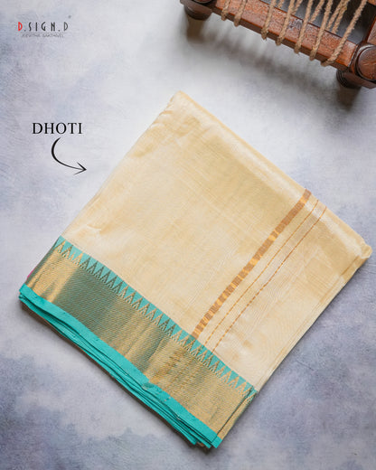 A folded light beige silk dhoti with a teal border and gold zari detailing, displayed on a textured gray surface with a wooden object partially visible in the top right corner.