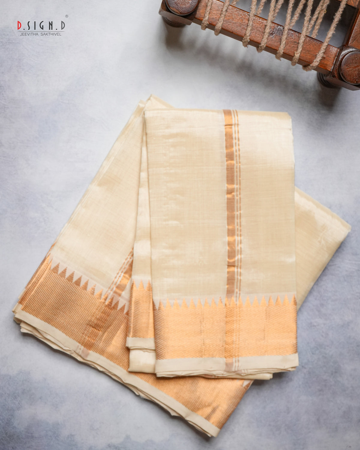 A folded cream-colored silk dhoti with a gold border and vertical stripes, displayed on a textured gray surface next to a wooden loom machine.