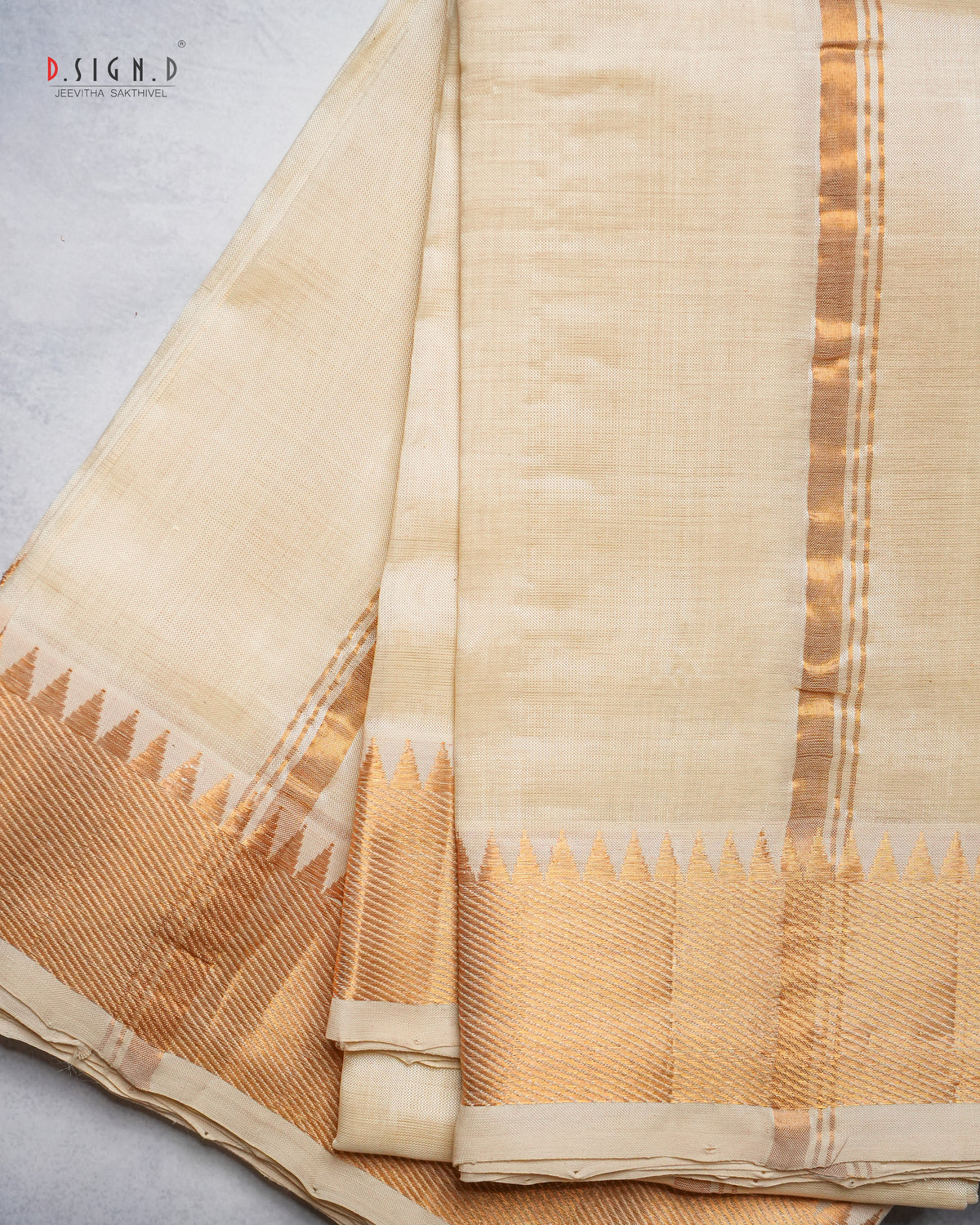 A folded cream-colored silk dhoti with a gold zari border and a decorative gold and beige pallu, displayed on a light gray textured surface.