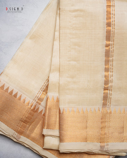A folded cream-colored silk dhoti with a gold zari border and a decorative gold and beige pallu, displayed on a light gray textured surface.