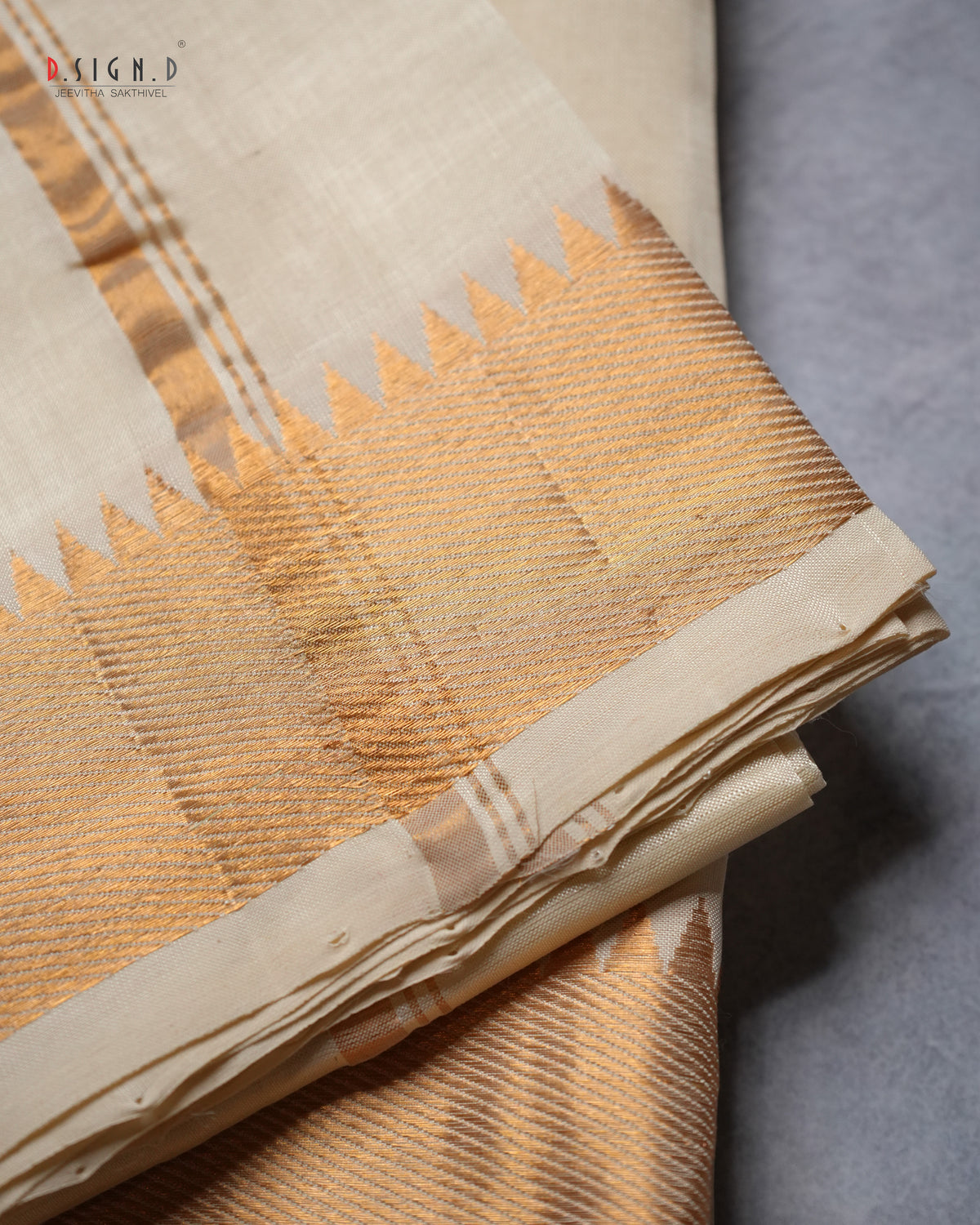A folded beige and gold silk dhoti with a decorative gold zari border, displayed on a neutral gray surface. 