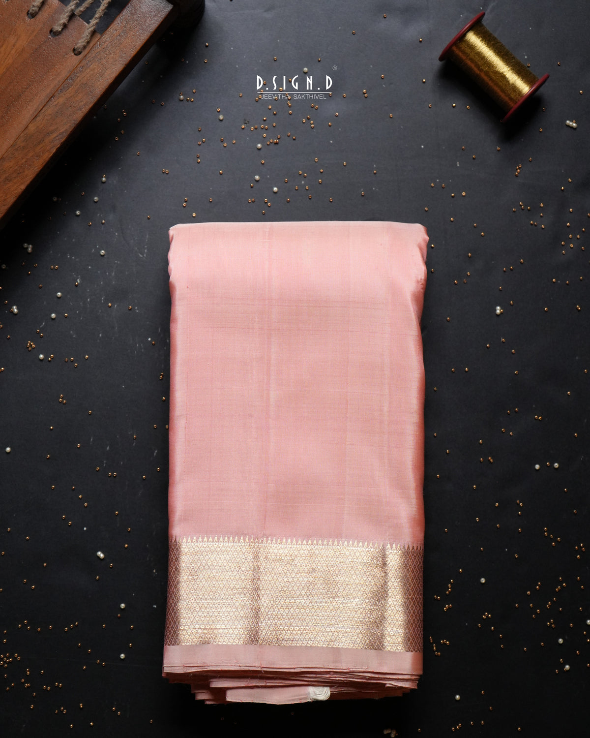 A folded dusty peach dhoti with a wide gold zari border, displayed on a dark textured background with decorative props.