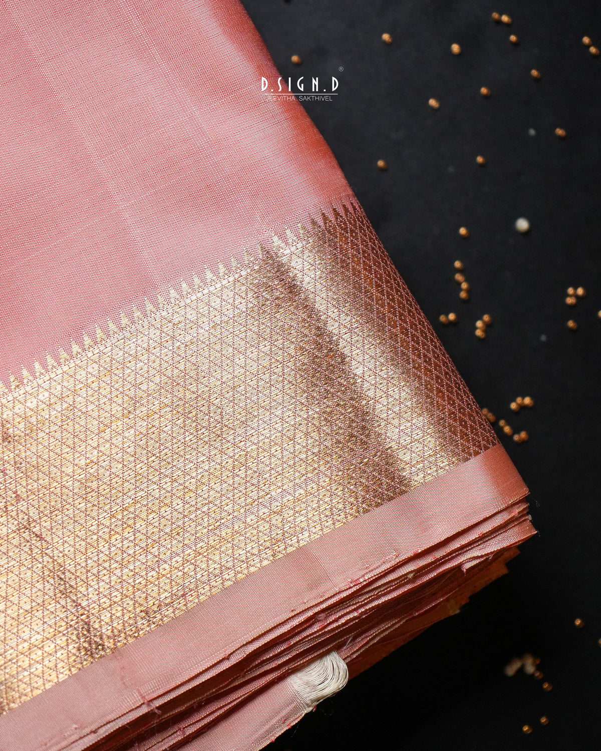 A folded dusty pink silk dhoti with an ornate gold zari brocade border, displayed on a dark textured background with scattered gold seeds. 