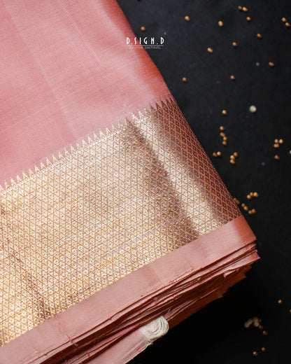 A folded dusty pink silk dhoti with an ornate gold zari brocade border, displayed on a dark textured background with scattered gold seeds. 