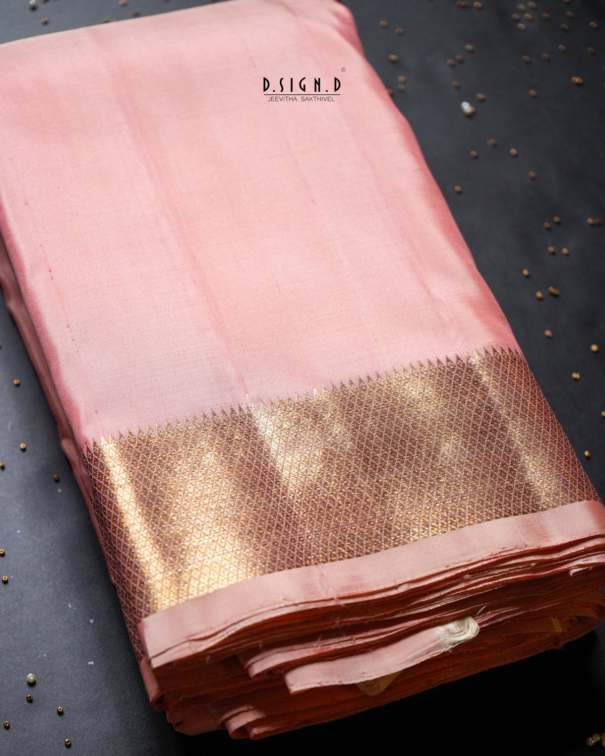 A folded pink silk dhoti with a wide metallic gold zari border, displayed on a dark textured surface with small scattered seeds.