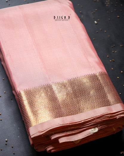 A folded pink silk dhoti with a wide metallic gold zari border, displayed on a dark textured surface with small scattered seeds.