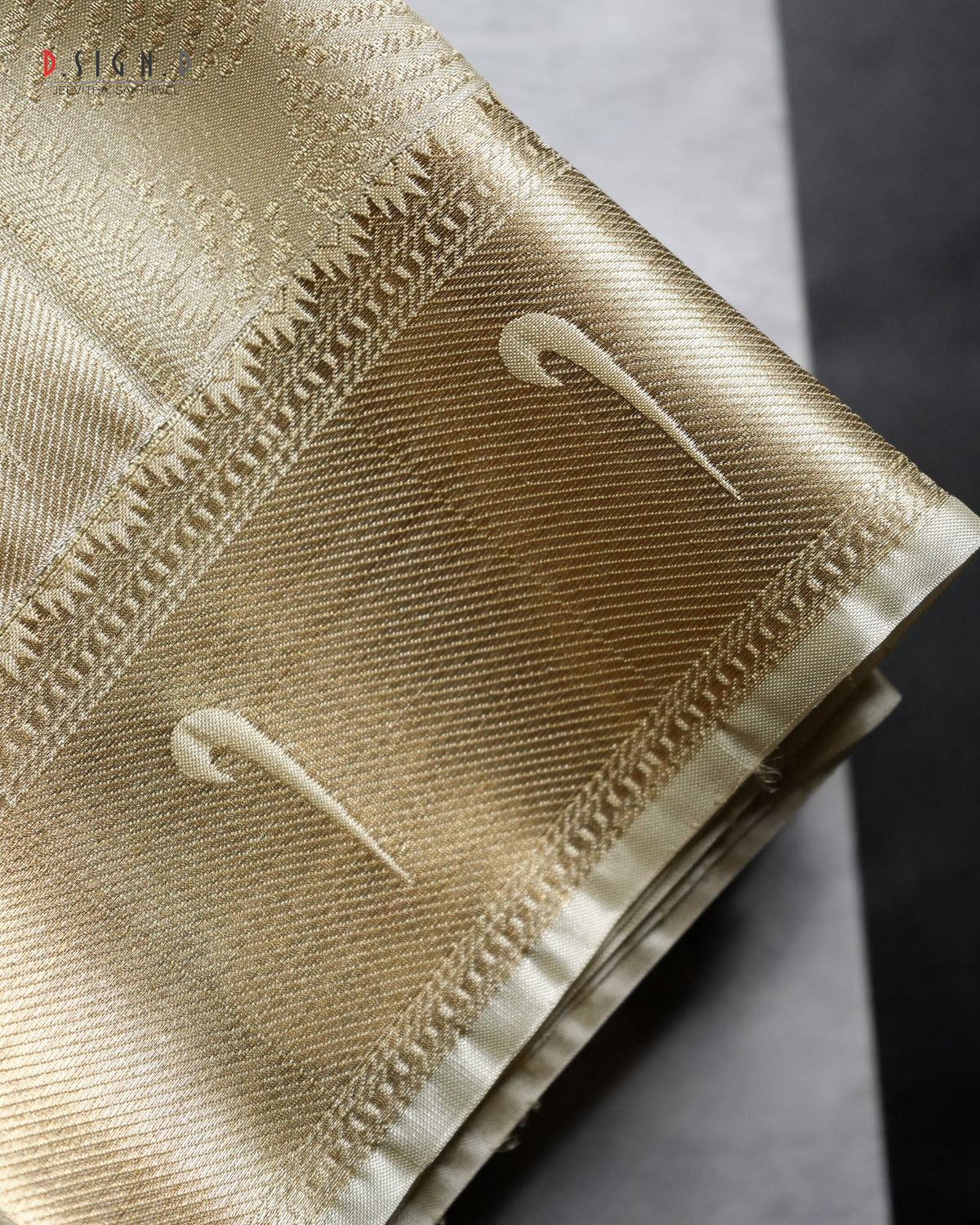 A close-up of a folded gold fabric with a woven pattern, featuring two stylized white swooshes and decorative stitched borders, set against a dark gray background.