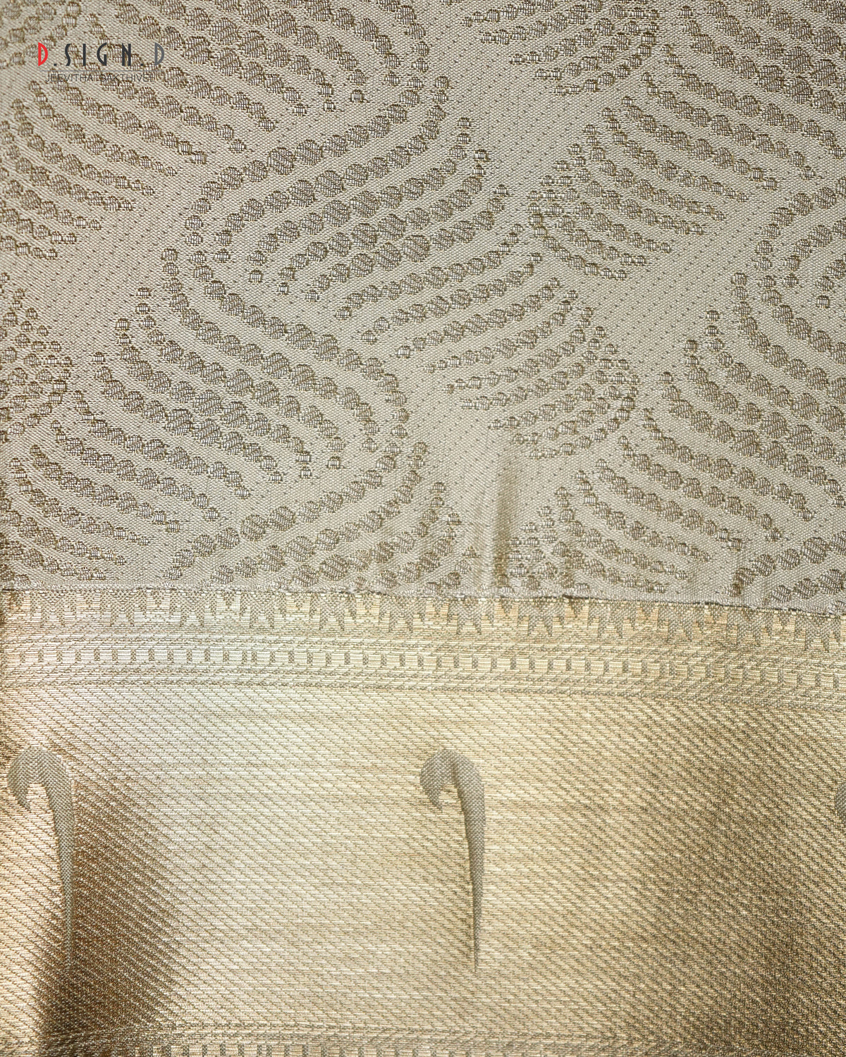 Close-up of a beige and gold patterned fabric, showing a textured upper section with repeating leaf-like motifs and a smoother decorative border below.
