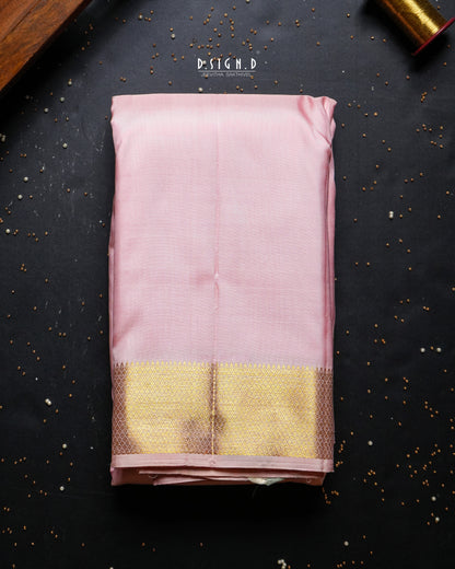A folded light pink dhoti set with a wide gold zari border, displayed flat on a dark textured background with decorative brass and wood elements.