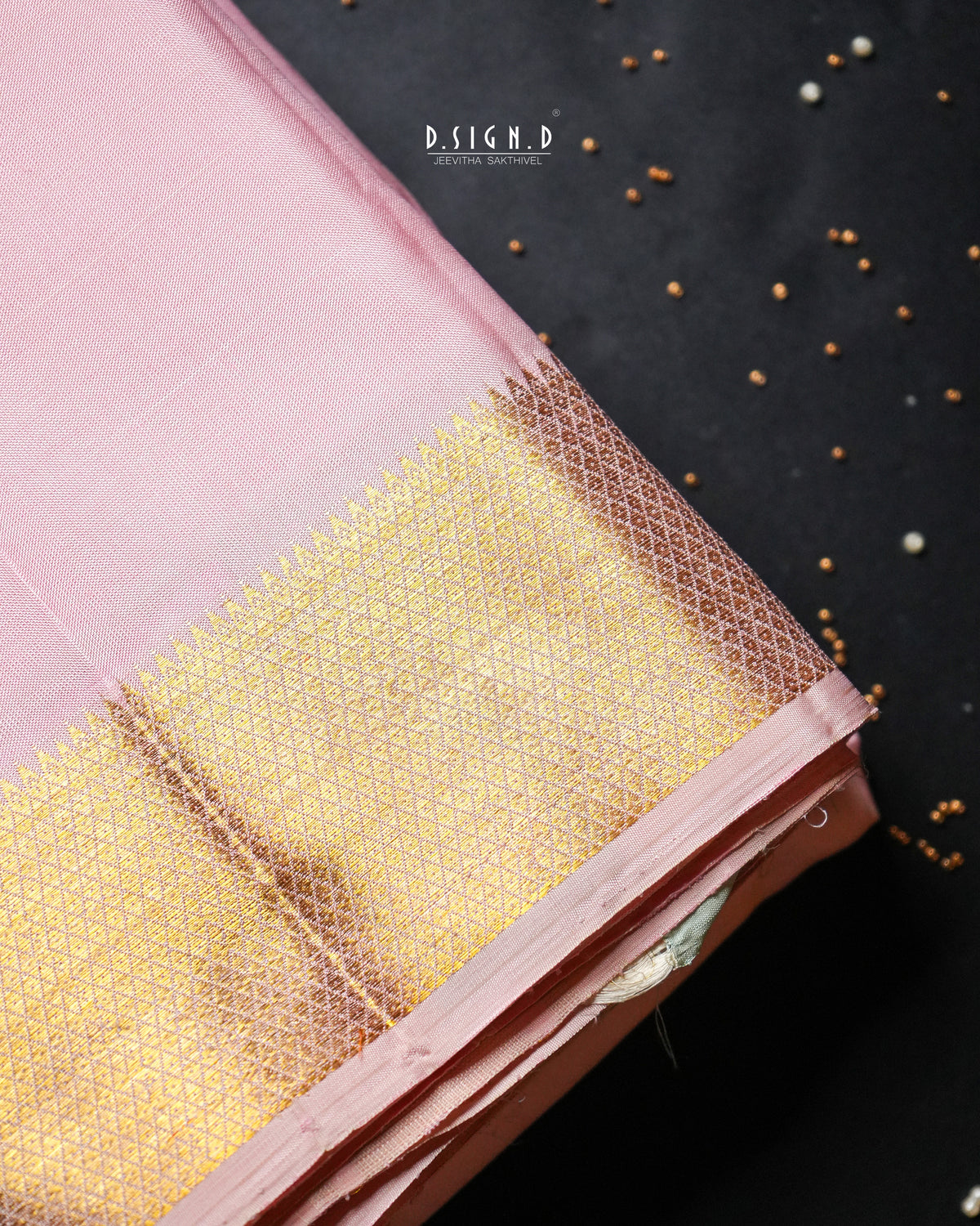 A close-up of a pink dhoti or fabric with a wide gold zari woven border, laid diagonally on a dark background with scattered small metallic beads. 