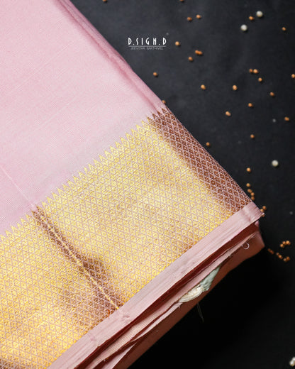 A close-up of a pink dhoti or fabric with a wide gold zari woven border, laid diagonally on a dark background with scattered small metallic beads. 