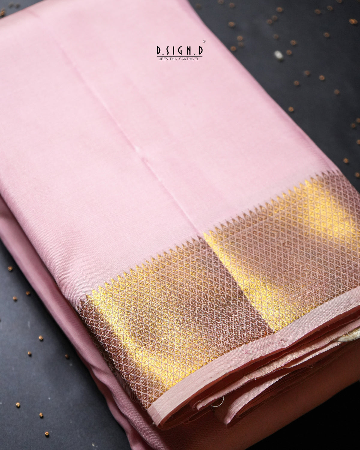 A folded pink fabric, likely a dhoti, featuring a wide metallic gold zari border with a woven geometric pattern, displayed on a dark textured background.