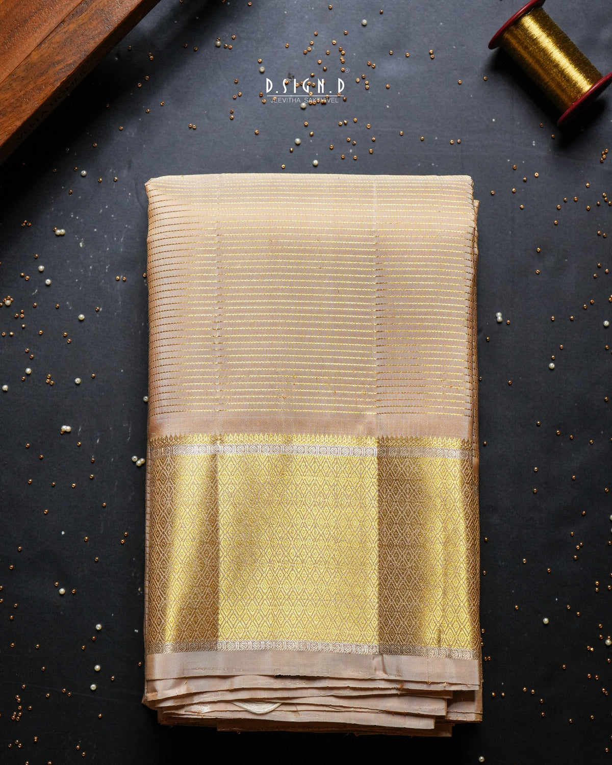 A folded beige and gold woven dhoti,  displayed on a dark textured background with decorative props including a spool of gold thread and scattered mustard seeds. 