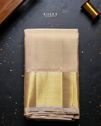A folded beige and gold woven dhoti,  displayed on a dark textured background with decorative props including a spool of gold thread and scattered mustard seeds. 