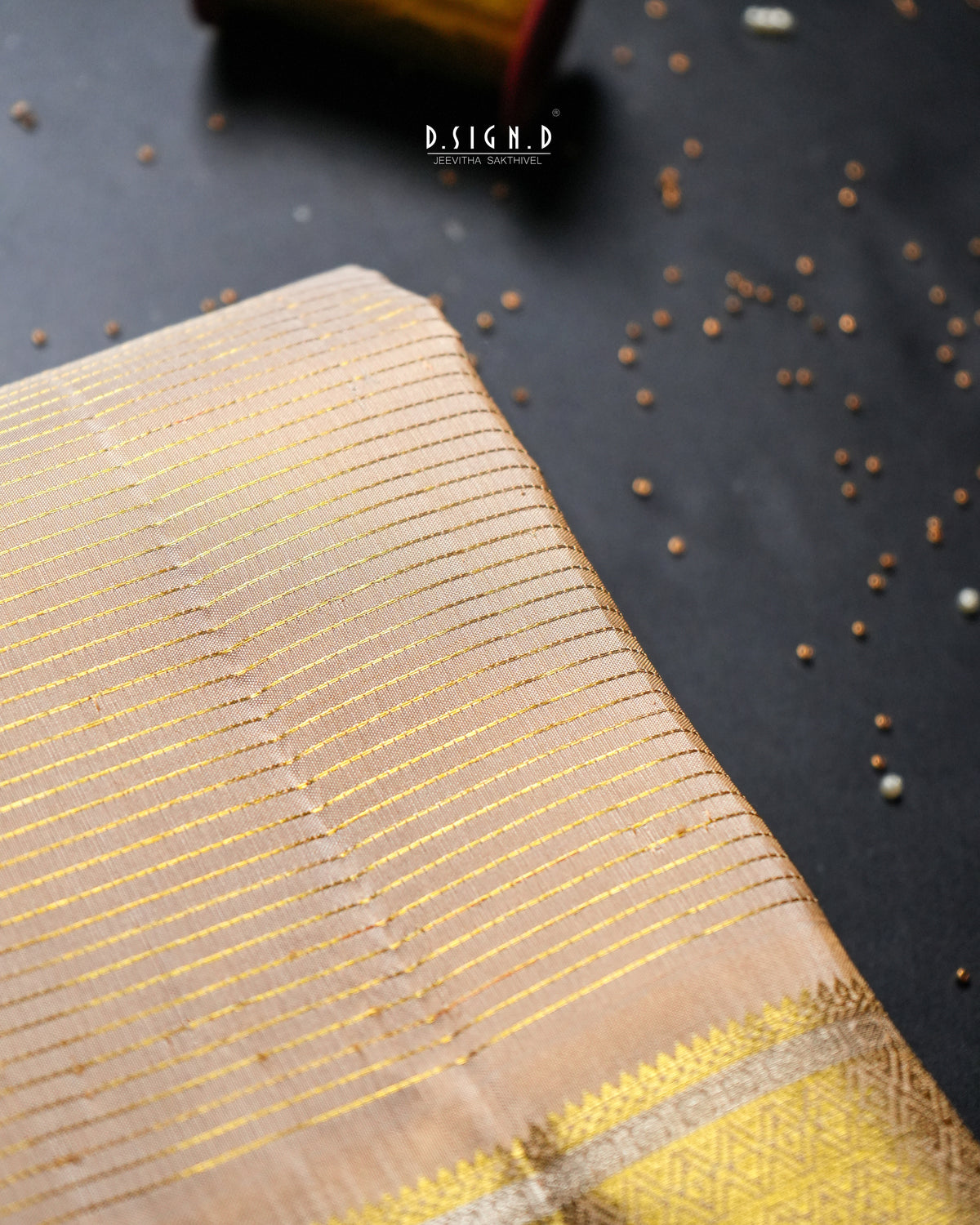 A close-up of a beige and gold striped dhoti, laid diagonally on a dark textured surface with scattered decorative seeds. 