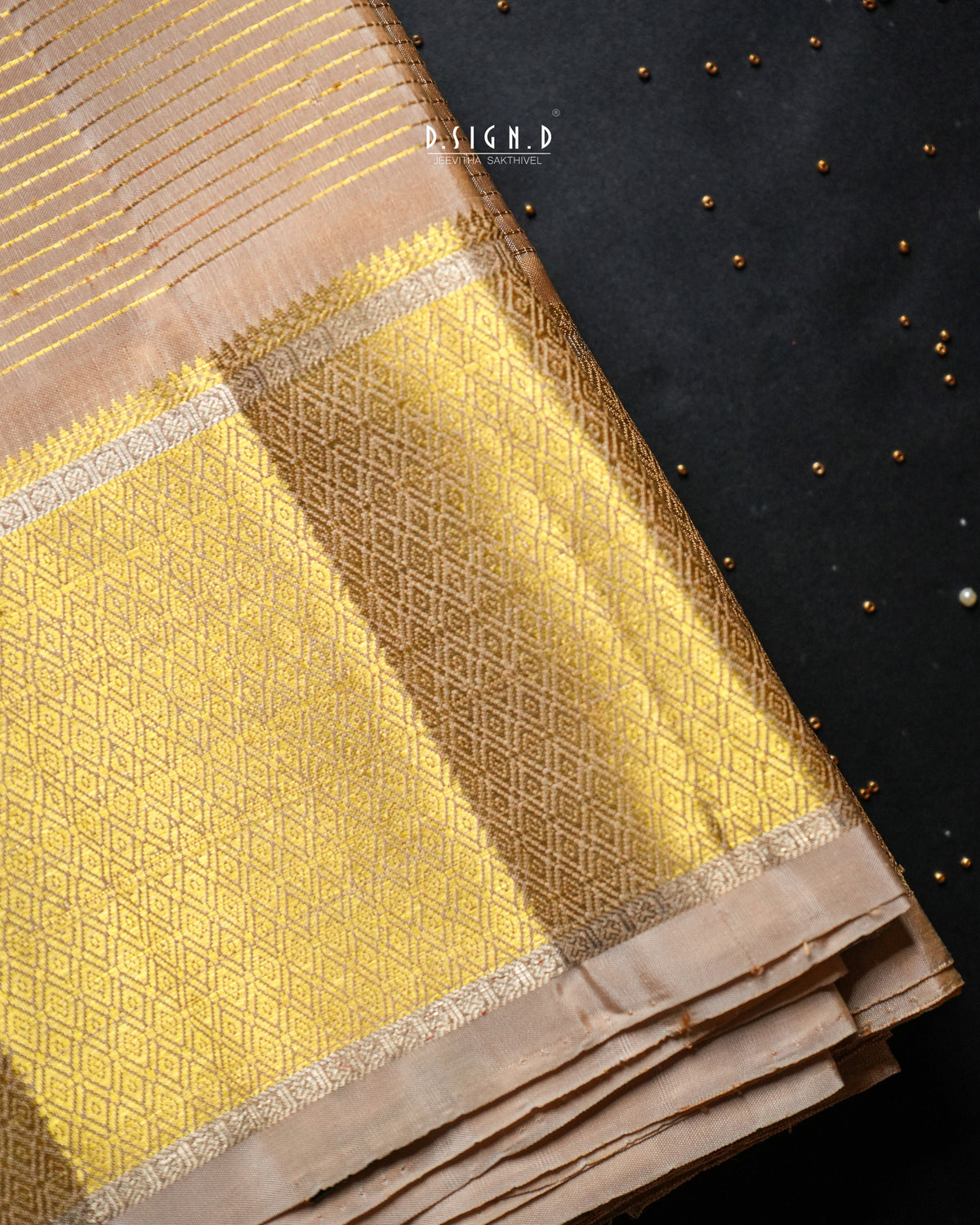 A folded gold and beige patterned dhoti, likely a saree or scarf, displayed diagonally on a dark textured background with small gold beads.