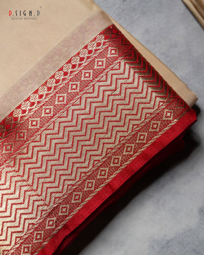 A close-up of a folded beige fabric with a wide red and beige patterned border featuring zigzag and diamond motifs, photographed on a gray surface.
