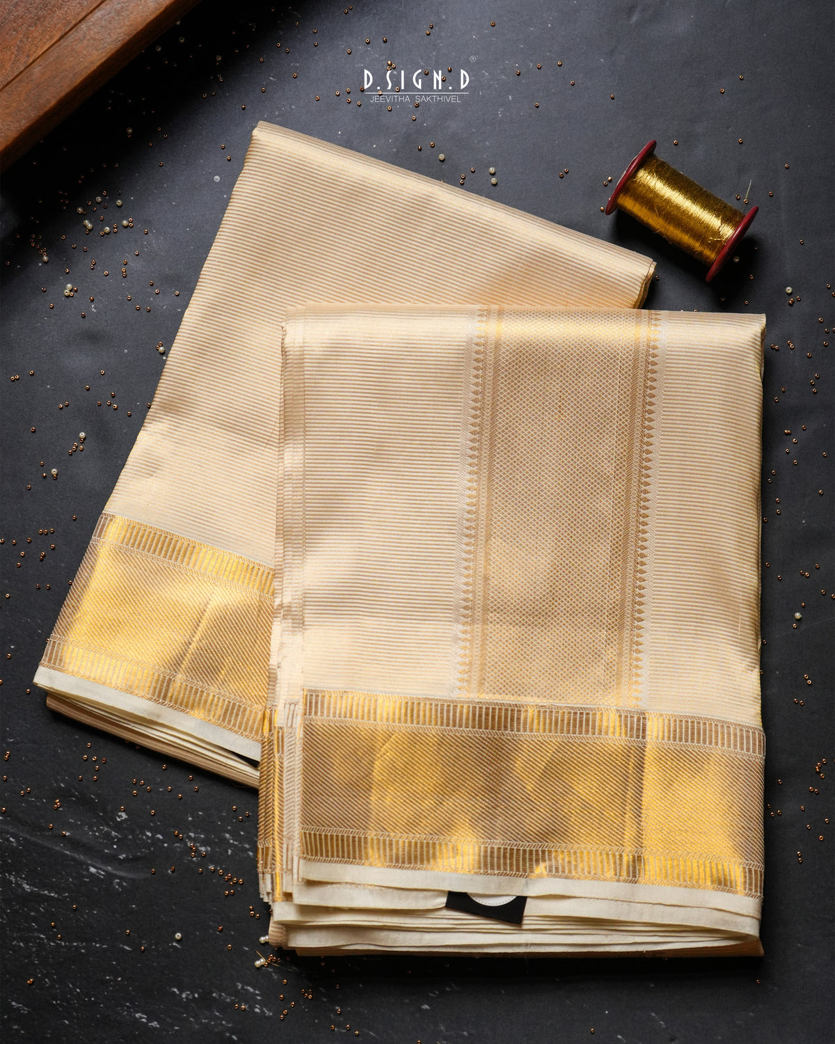 Two folded beige silk dhoti & vastram with wide metallic gold borders are arranged on a dark textured surface, with a spool of gold thread and a small wooden block as styled props. 
