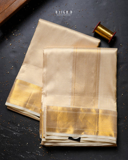 Two folded beige silk dhoti & vastram with wide metallic gold borders are arranged on a dark textured surface, with a spool of gold thread and a small wooden block as styled props. 