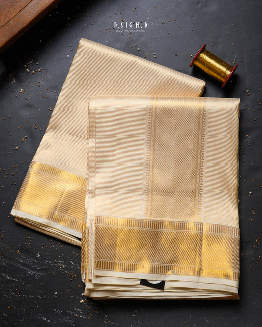 Two folded beige silk dhoti & vastram with wide metallic gold borders are arranged on a dark textured surface, with a spool of gold thread and a small wooden block as styled props. 