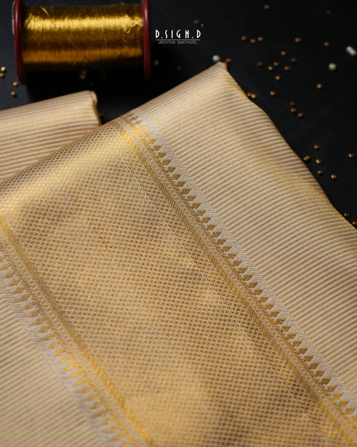 A close-up of a folded beige fabric with a fine woven texture and a decorative zigzag border, shown on a dark background with a spool of gold thread in the corner. 