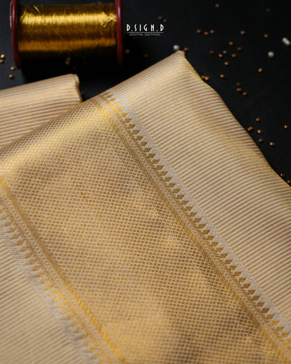 A close-up of a folded beige fabric with a fine woven texture and a decorative zigzag border, shown on a dark background with a spool of gold thread in the corner. 