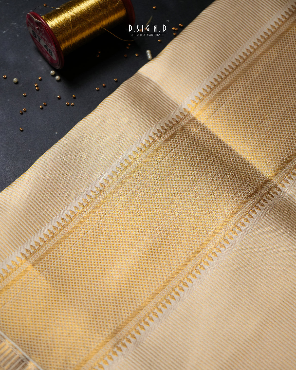 A close-up of a beige woven textile with a subtle dotted pattern and decorative border, laid diagonally on a dark surface with a gold spool prop in the corner. 