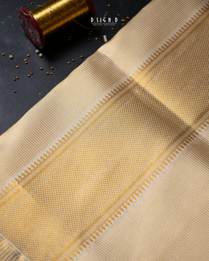 A close-up of a beige woven textile with a subtle dotted pattern and decorative border, laid diagonally on a dark surface with a gold spool prop in the corner. 